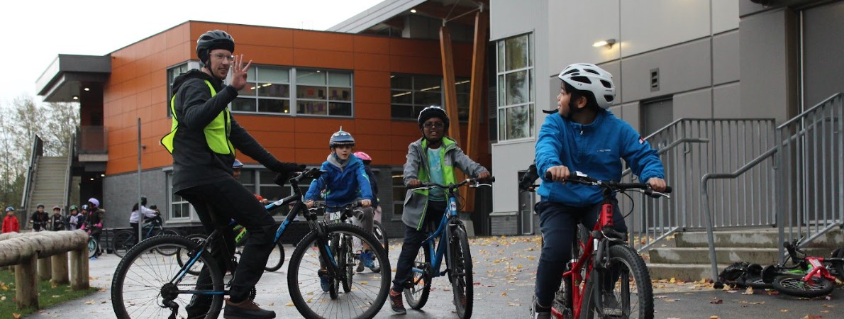A school-aged student on a bike shoulder checks. An instructor behind them holds up four fingers.