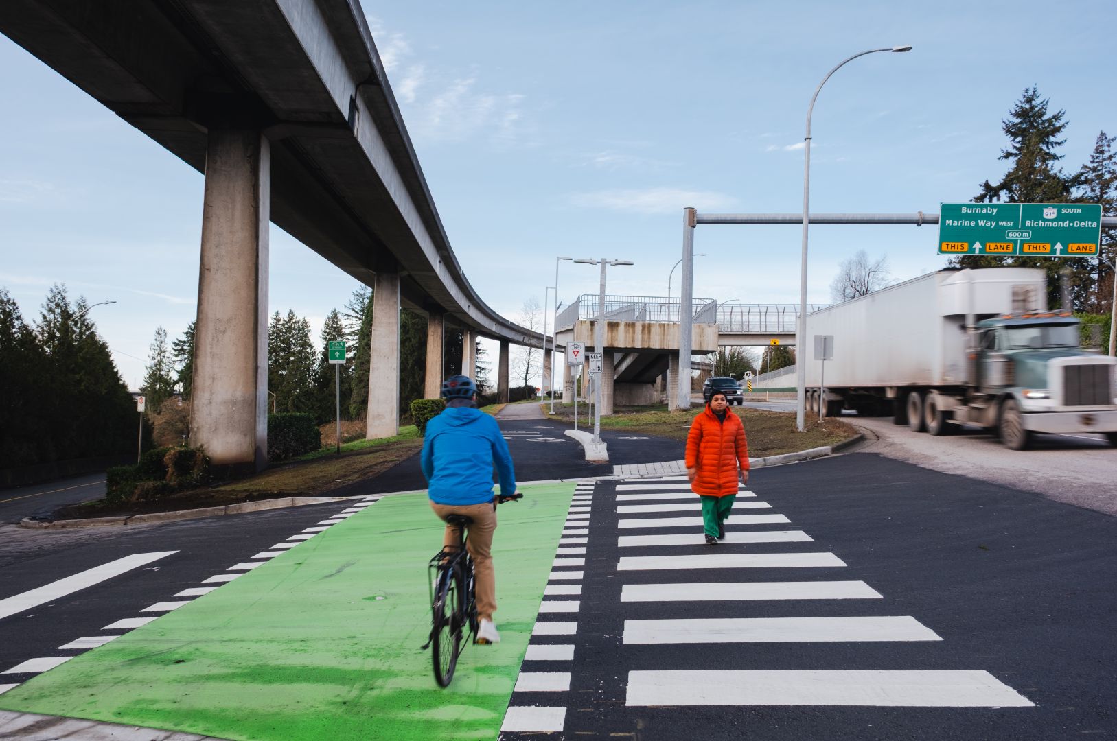 A cyclist in a blue jacket rides on a bright green bike lane, approaching a crosswalk where a person in an orange coat is walking. The scene is set beneath an elevated SkyTrain track with traffic signs and trucks in the background.