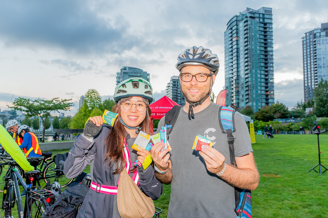 A woman on the left and a man on the right wearing bike helmets at David Lam Park during Bike the Night holding snacks from Herbaland, Organika, Ritter Sport, and Ener C