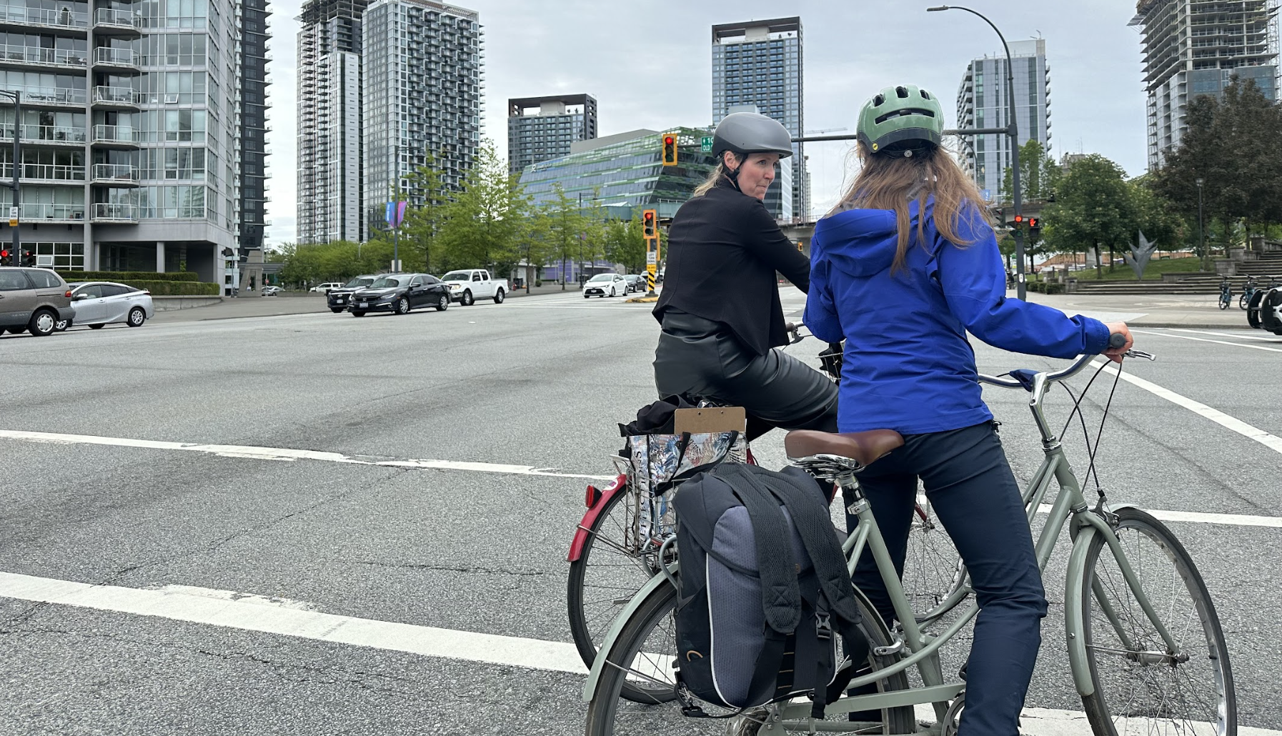 Two people wearing helmets on bicycles stopped at a multi-lane city intersection, with modern high-rise buildings in the background.