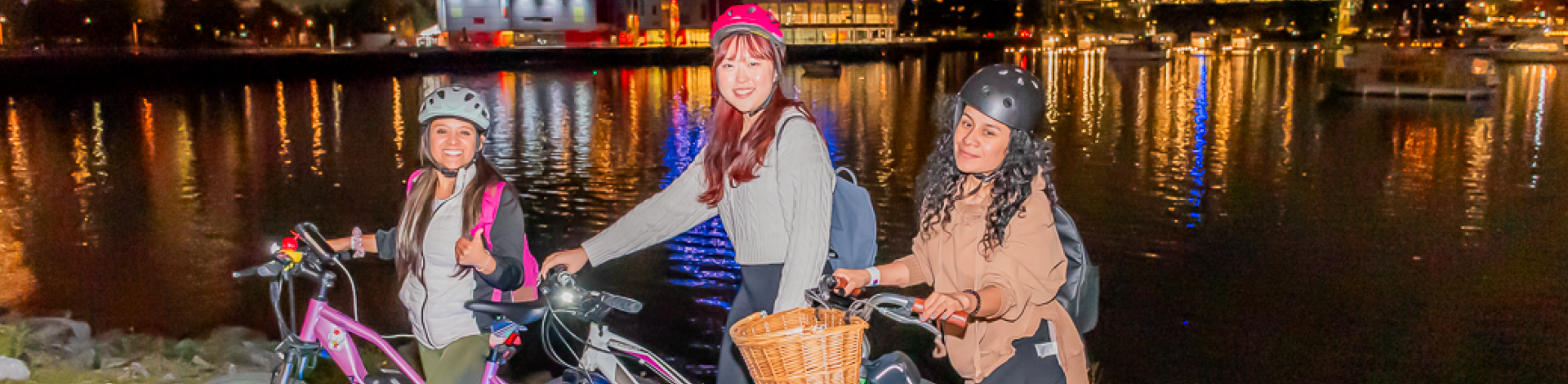 Three women stand with their bikes along Vancouver's Seawall. It is night time.