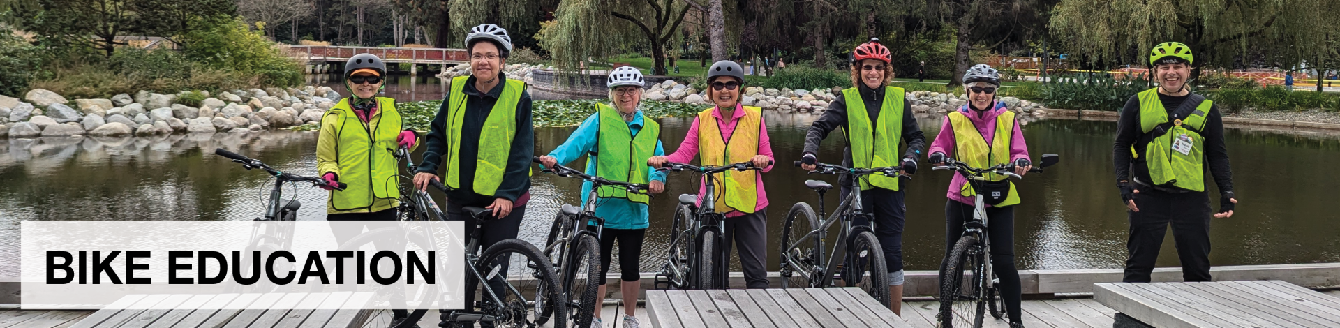 A group of six participants and one bike education instructor pose with their bikes on a wooden path. There is a pond of water behind them. They are all wearing yellow vests.