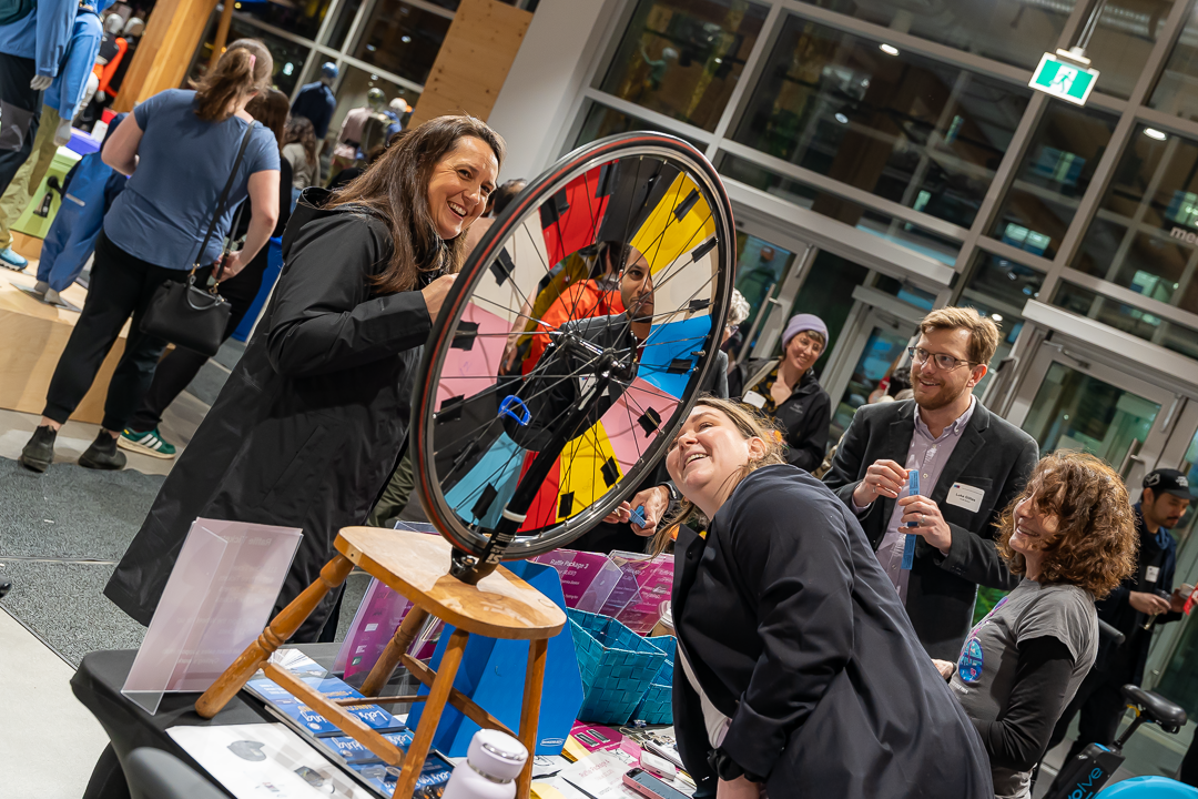 A group of people gathers around a colorful prize wheel made from a bicycle wheel at an indoor event. One woman spins the wheel while others watch and laugh, clearly enjoying the activity. The table is filled with promotional materials, prizes, and signage. The setting appears to be a brightly lit retail store with glass doors and windows in the background.