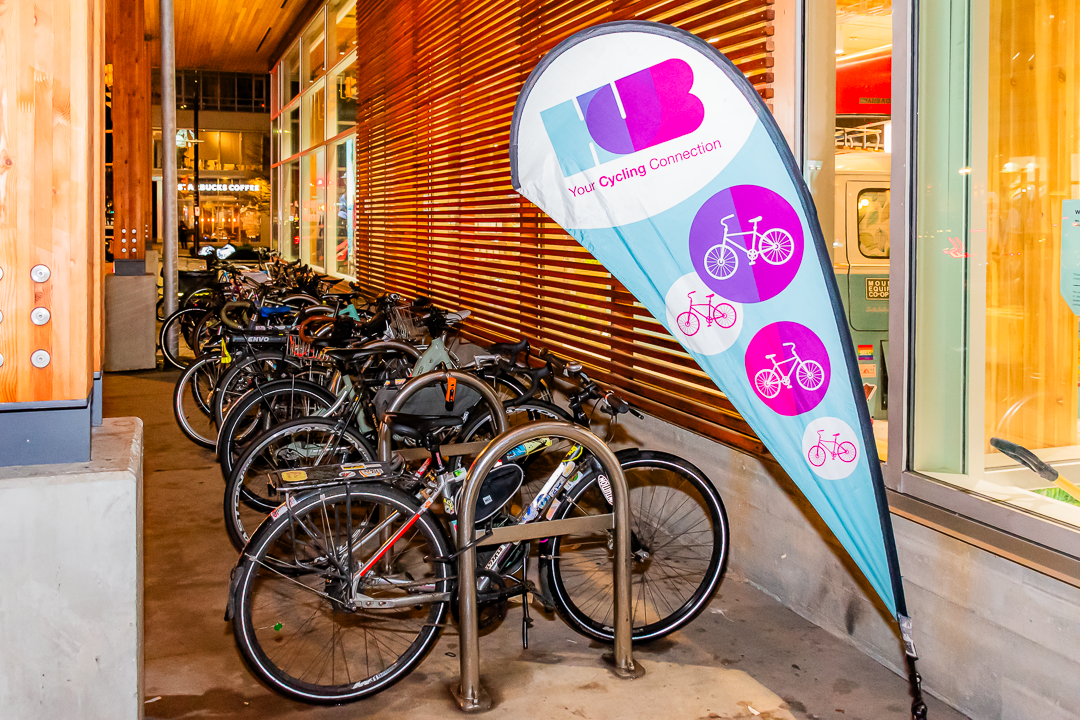 A row of bicycles is parked at a bike rack outside a building during the evening. A bright and colorful HUB Cycling banner reading "Your Cycling Connection" is prominently displayed next to the bikes. The scene is well-lit.