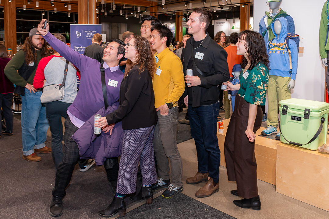 A group of six people pose for a cheerful selfie at an indoor event, smiling and holding drinks. The group stands in front of a retail display featuring mannequins dressed in outdoor clothing and a light green YETI cooler. Other attendees and event signage are visible in the background, creating a lively and social atmosphere.