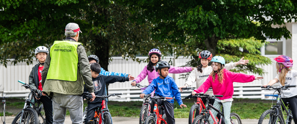 An instructor speaks to a group of students with their bikes. All of the students are indicating a left turn with their arm.