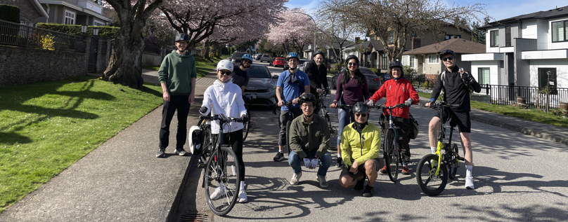 A group of ten cyclists pose on a residential street lined with blooming cherry blossom trees under a bright blue sky. Some are standing with their bikes, while others kneel or sit on their bicycles. The scene is sunny and cheerful, with houses and well-kept lawns visible on both sides of the street.