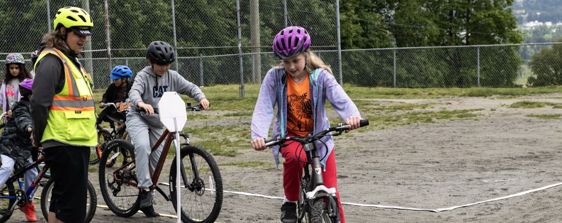 A child rides their bike on school grounds as a bike education instructor watches and gives notes.