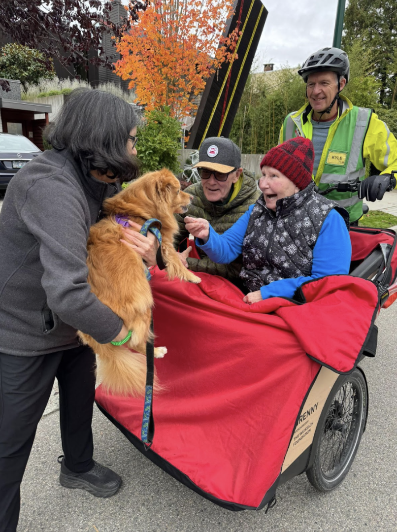 Two seniors being pedaled in a cycling without age trishaw are smiling as someone is hodling a dog eye level.