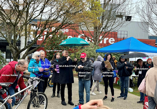 A group of people gather outdoors at a community event near the Maple Ridge Leisure Centre. Several individuals are labeled by name, including Michael Nelson, Jackie Chow, Ivan Chow, Sigrun Gilmour, MLA Bob D’Eith (holding a green umbrella), Mayor Dan Ruimy (wearing a red hat), Ashley van der Pauw, and Kirk Grayson. Tents and booths are set up in the background, and a person in the foreground is holding a bicycle. The atmosphere appears cheerful and community-focused.