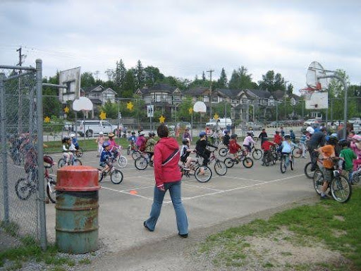 A large group of children ride bicycles around a fenced outdoor basketball court during what appears to be a cycling class or event. An adult in a red jacket stands in the foreground, overseeing the activity. Several basketball hoops are visible, and spectators or parents stand along the fence on the right. The background includes suburban houses and trees under a cloudy sky.