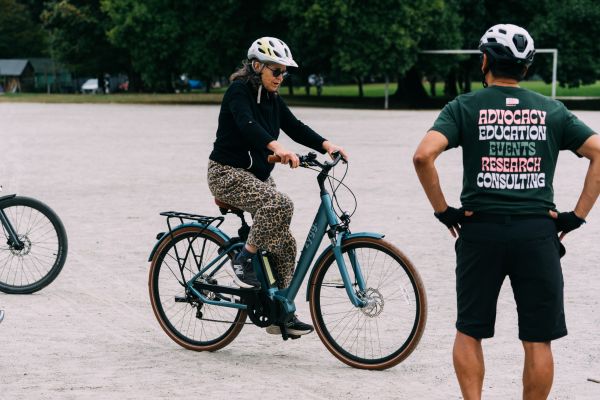A woman rides an e-bike on a gravel field while an instructor watches her.