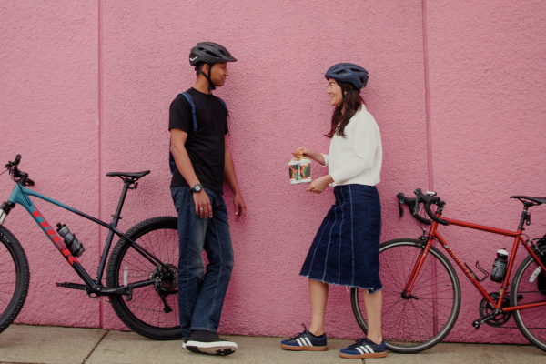 Two people stand against a pink wall with their bikes behind them. One woman is holding a 4-pack of beer.