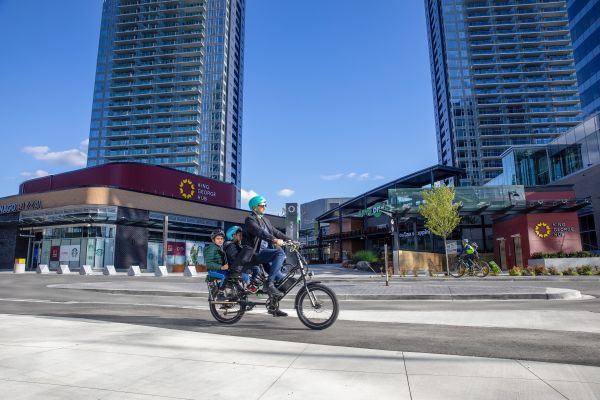 A person rides an electric cargo bike with two children as passengers in front of tall modern high-rise buildings and shops at King George Hub. The sky is clear and blue, and everyone is wearing teal helmets. The scene is urban, with contemporary architecture and people walking or cycling nearby.