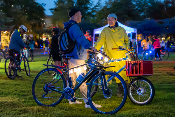 Two people laugh and chat with one another at HUB Cycling's Bike the Night event. They are both standing with their bikes. There are several other people with bikes in the field behind them.