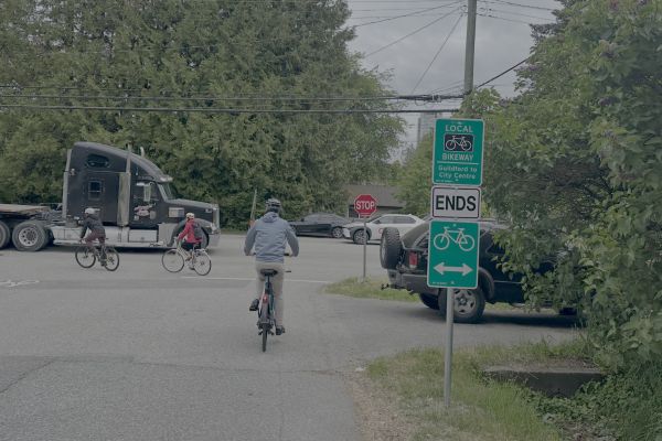 A group of three cyclists, all wearing helmets, ride through a quiet street intersection, with a large black semi-trailer truck and several cars nearby. Prominent green bikeway signs indicate that the local bikeway ends here and direct cyclists to Guildford or the city centre, while another sign points left and right for bike routes. Trees and bushes line the roadway, and the area appears overcast.