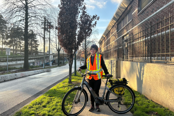 Chelsea standing on the sidewalk with an orange reflective vest and with the ohm e-bike.
