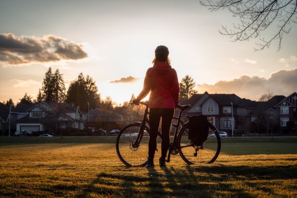 A woman stands in a field with her bike. She is staring off into the distance at the sunrise.