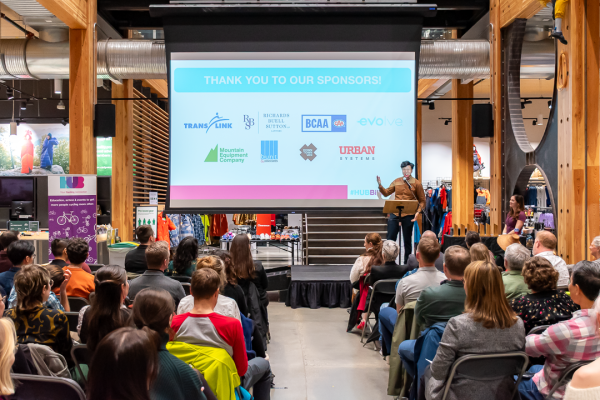 A speaker stands on stage at a HUB Cycling event, addressing a seated audience in a retail store setting. A large screen behind them displays a "Thank You to Our Sponsors!" slide with logos of organizations including TransLink, Mountain Equipment Company, BCAA, Urban Systems, and others. Attendees are attentively watching the presentation. The venue features wood and metal architectural details, and outdoor gear is visible in the background.
