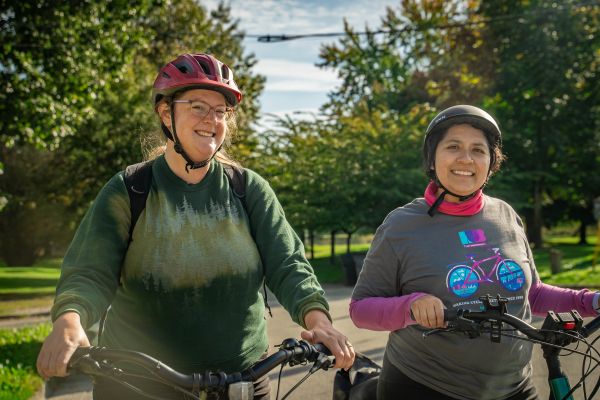 Two women smile as they stand with their bikes. Both women are wearing helmets.