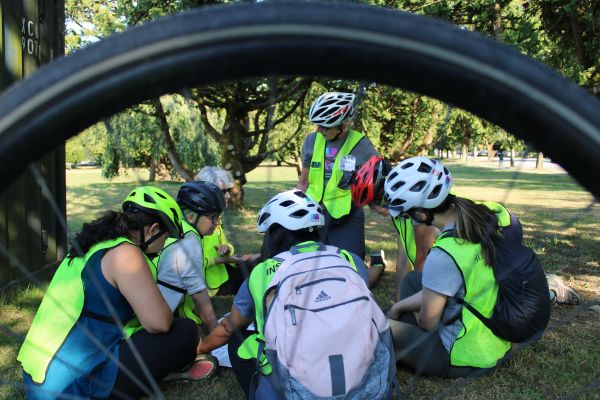 Looking through the spokes of a bike wheel, HUB instructor is giving a lesson with participants sitting on the grass reviewing course material