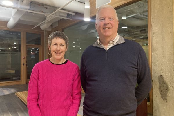 Victoria Gray and Jeff Leigh stand side by side. Victoria is a middle-aged woman wearing a bright pick sweater. Jeff Leigh is a middle-aged man wearing a black sweater.