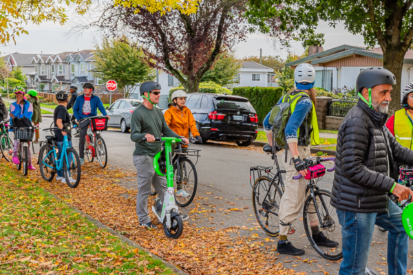 group of people on scooters and bikes waiting on a residential street with colorful leaves around them