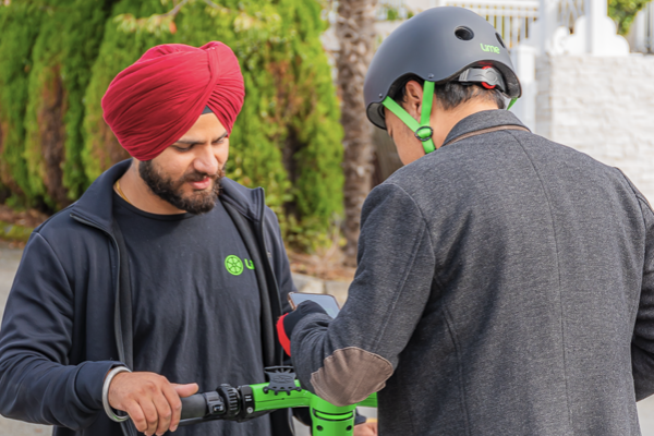 A man wearing a turban stands on a Lime scooter while another man wearing a helmet helps him activate the scooter.