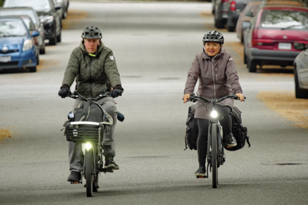 A man and a woman ride their bikes side by side on a residential road. Cars are parked on both sides of the road.
