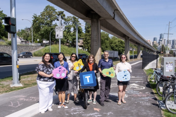 A group of seven people stand smiling with colorful signs shaped like bicycle silhouettes under an elevated SkyTrain track. They are gathered on a multi-use path marked with chalk messages and a "Welcome to BC Parkway" sign.