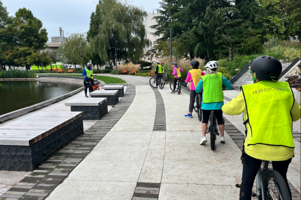 Several people ride their bikes on a multi-use path. They are wearing yellow pinnies.