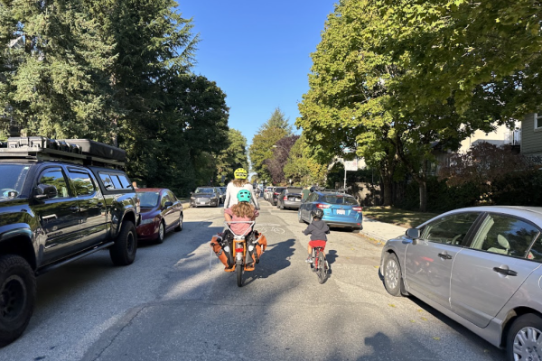 A mother and her two children ride their bikes on a residential roadway in Vancouver. Cars are parked on both sides of the street.