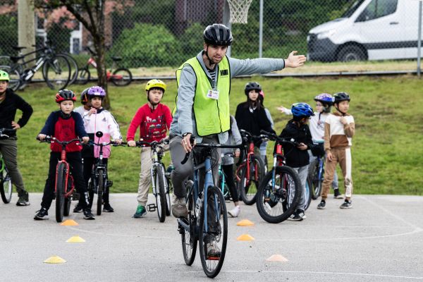 A bike education instructor shows how to make a left-hand turn signal. Several students on bikes watch.