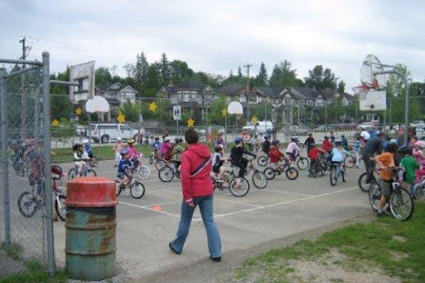 A large group of children ride bicycles around a fenced outdoor basketball court during what appears to be a cycling class or event. An adult in a red jacket stands in the foreground, overseeing the activity. Several basketball hoops are visible, and spectators or parents stand along the fence on the right. The background includes suburban houses and trees under a cloudy sky.