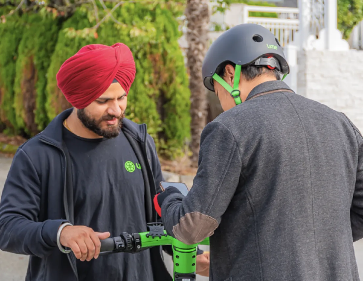 A man wearing a turban stands on a Lime scooter while another man wearing a helmet helps him activate the scooter.