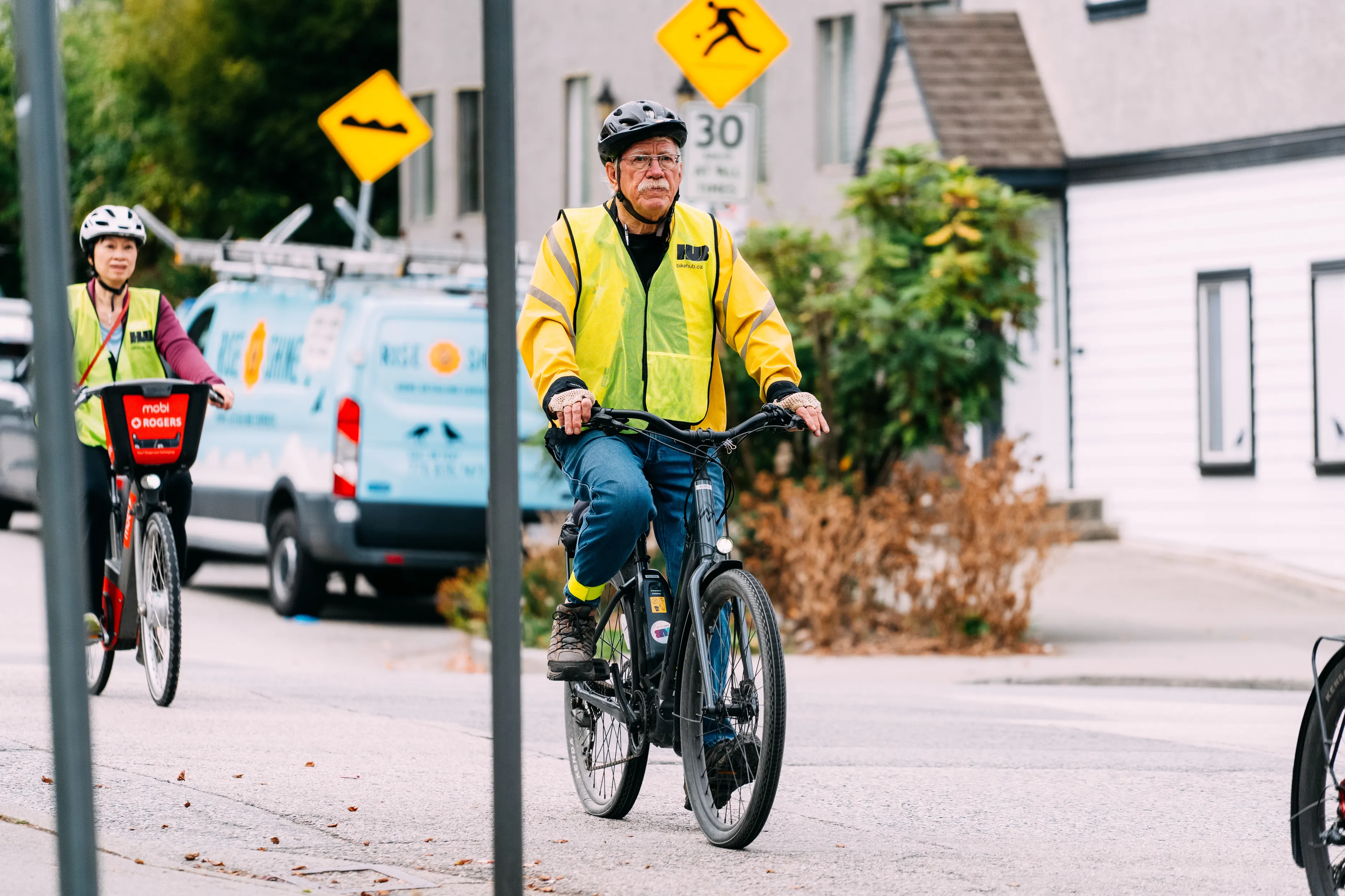 Two people ride their bikes on the road. They are both wearing yellow HUB jerseys.