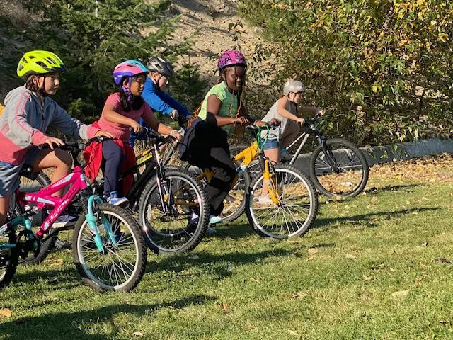 Several school children ride their bikes in a race-like fashion on grass.
