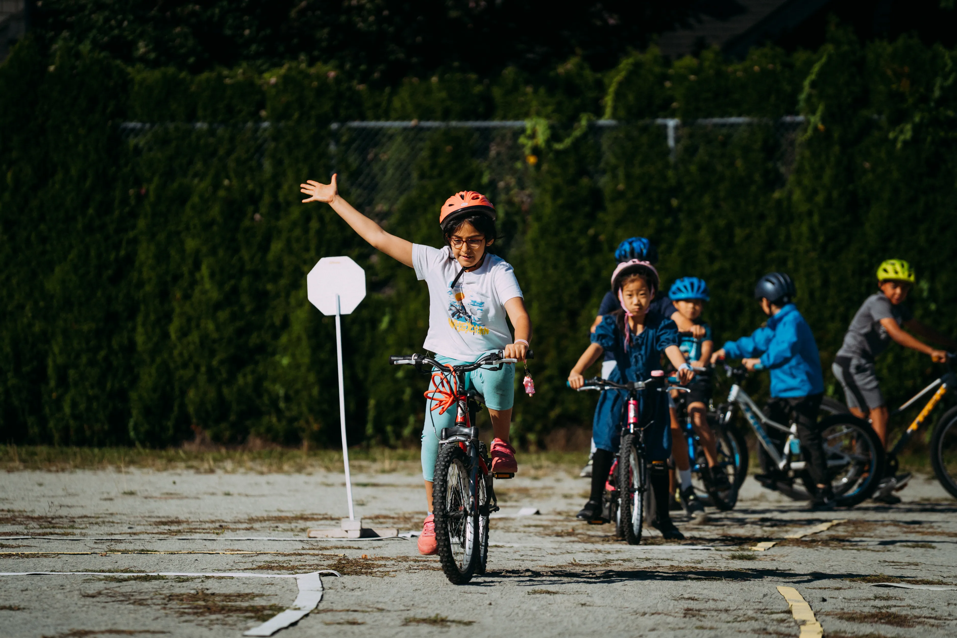 A elementary-school aged girl rides a bike on school grounds and indicates a right-turn signal with her arm. Several students are waiting behind her with their bikes.