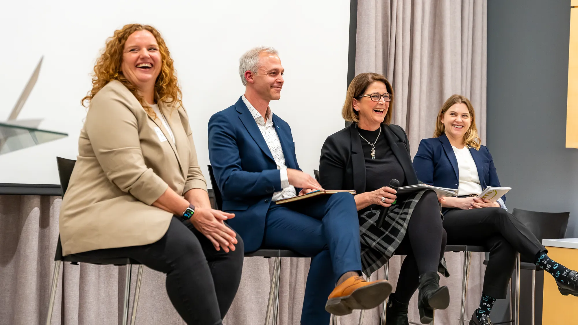 Four panelists laugh. All four are seated in a row of seats on stage.
