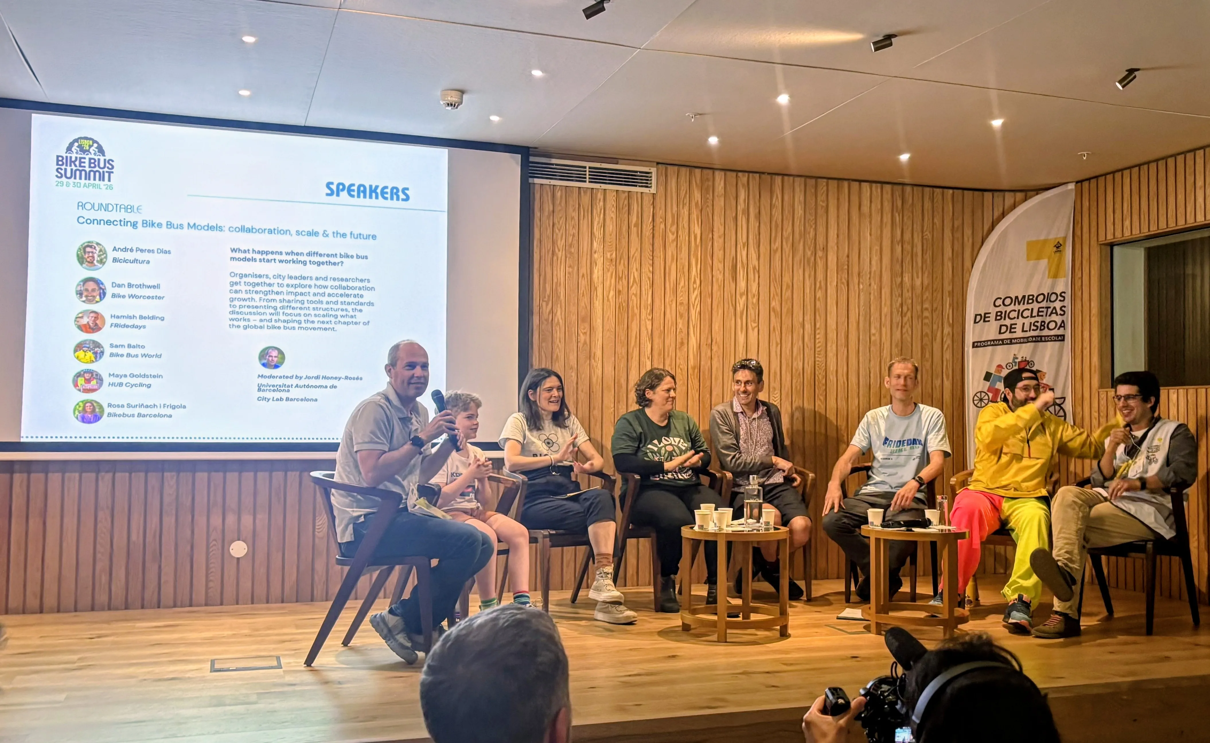 A group of people sit in a row on stage and discuss. There is a PowerPoint presentation playing on a screen behind them.