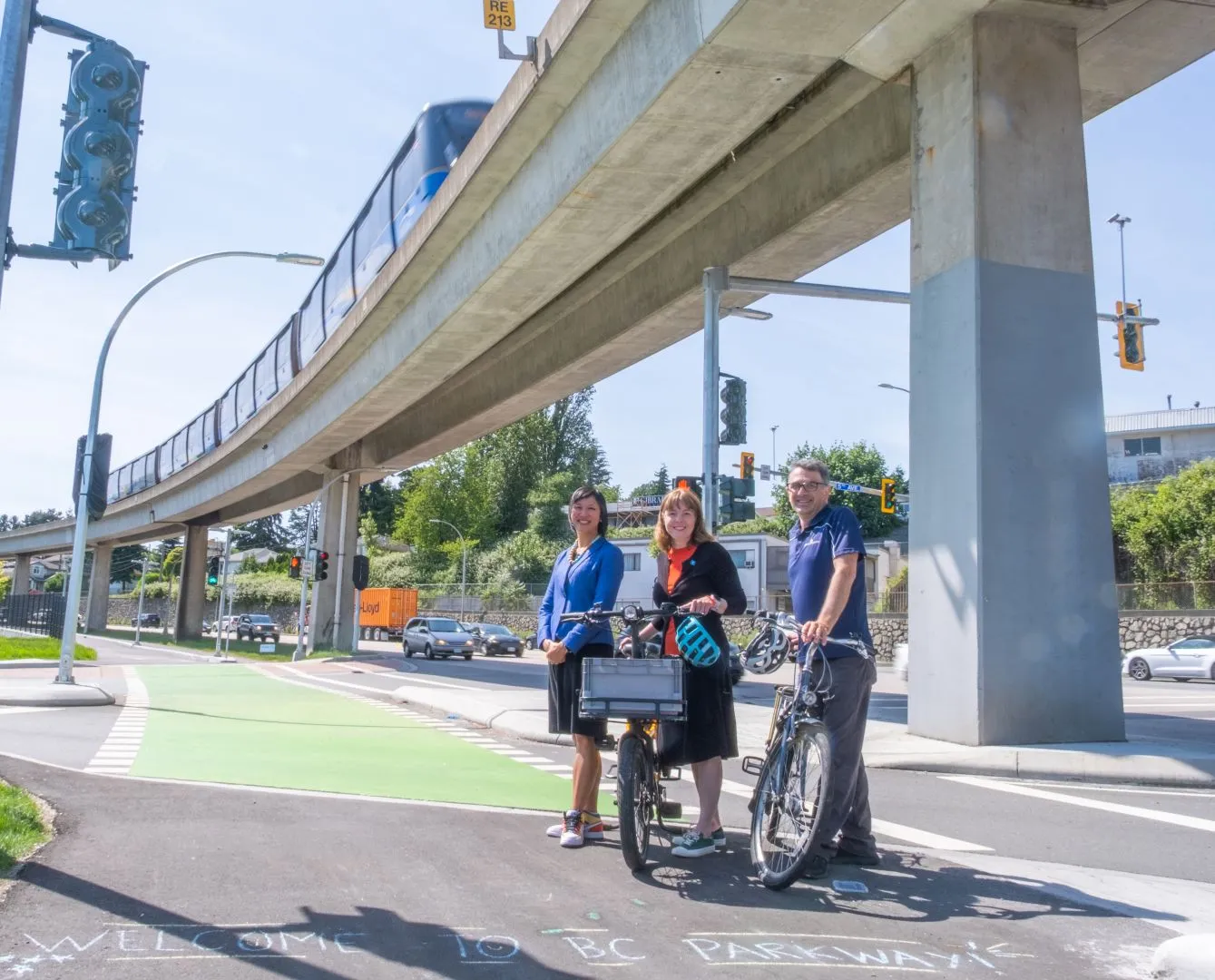 Three people stand with their bikes at the new Stewardson Way cycling infrastructure. The SkyTrain line is above them.