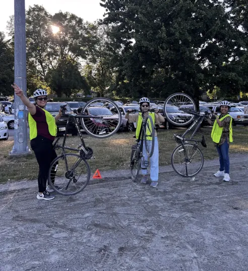 Three adult women hold their bikes up on one wheel. They are all wearing yellow pinnies.
